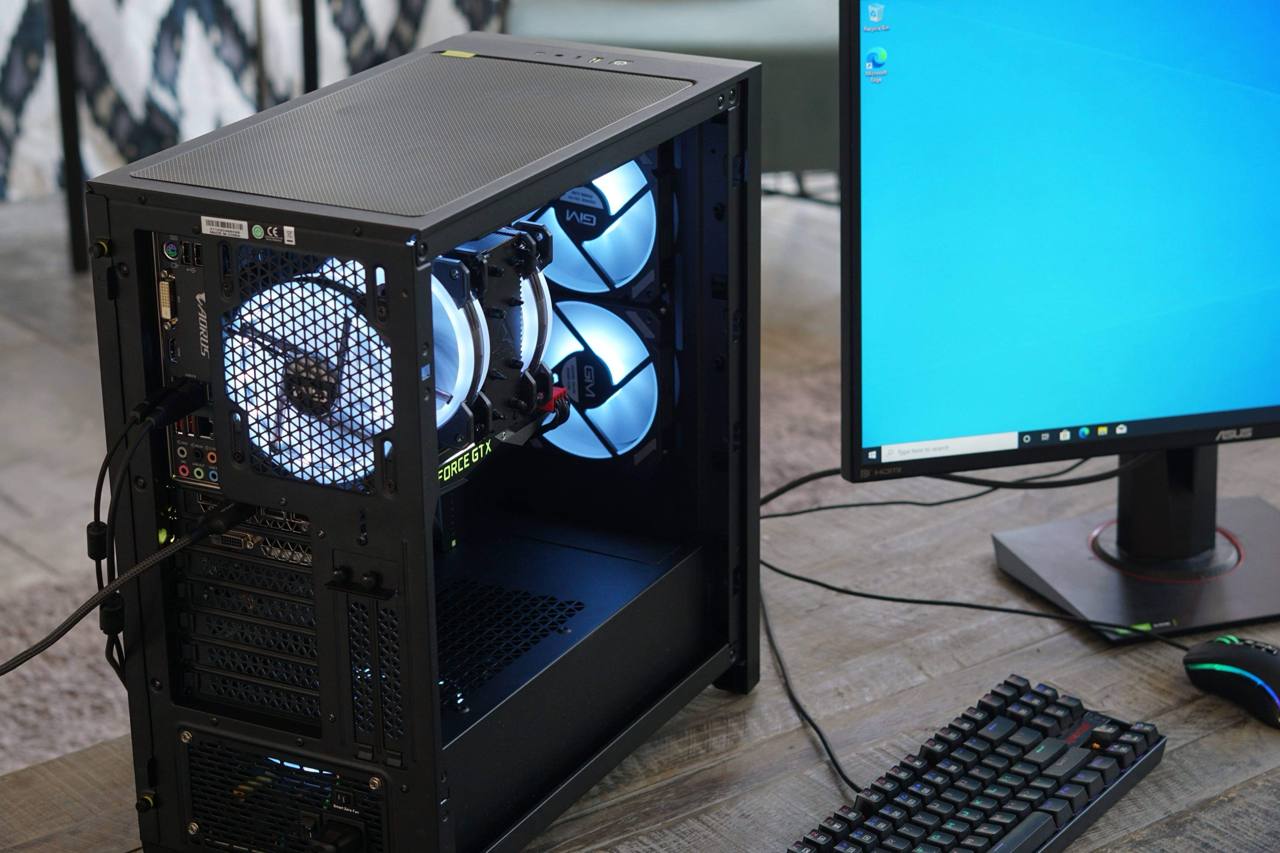 Black PC viewed from the rear side with white-blue fans beside a monitor and keyboard